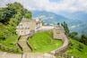 Un château en pierre doté d’un escalier en colimaçon en bois, entouré d’une végétation luxuriante et avec des montagnes en toile de fond.