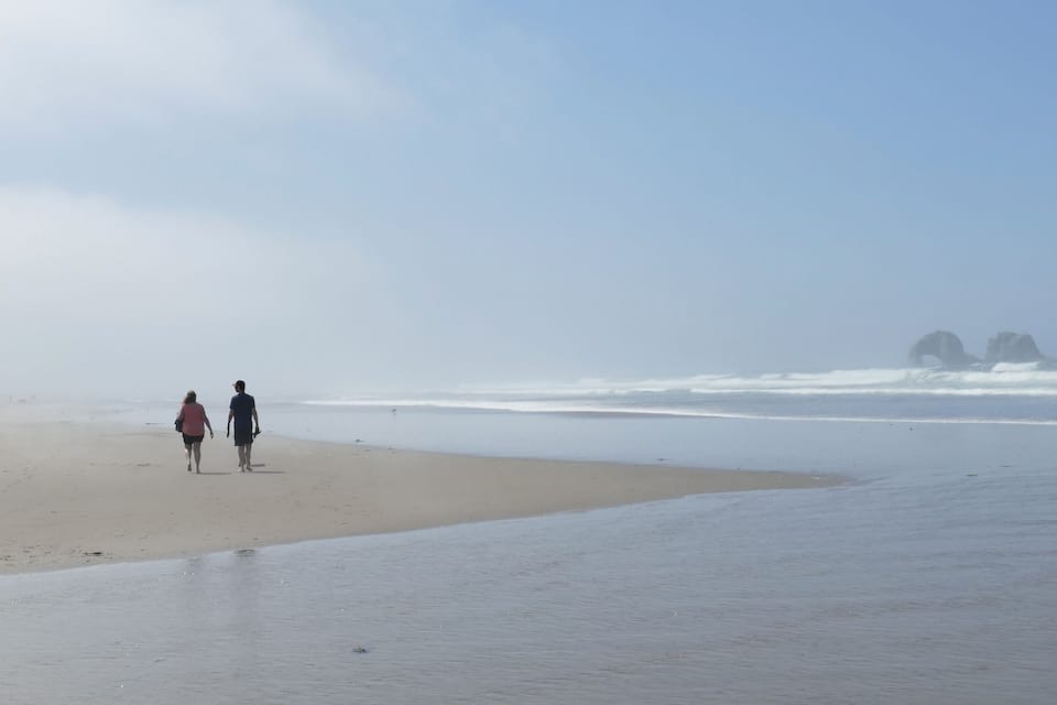 A beach with two people walking along the shore, waves crashing onto the sand, and distant rocky formations.