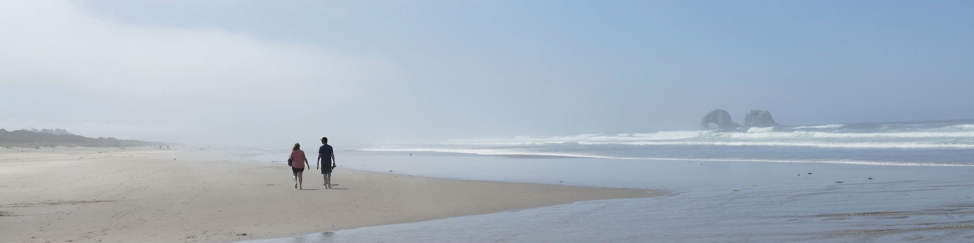 Misty Beach Walk Panorama- A couple walks the Rockaway Oregon beach in the morning before the mist has burn off. Twin Rocks sea stacks sit on the horizon.