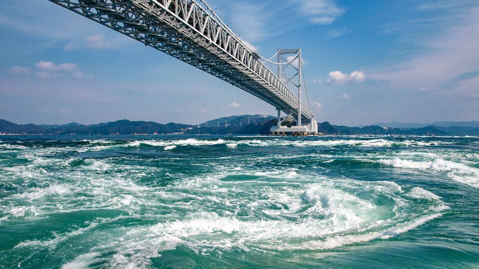 A large suspension bridge over turbulent ocean waters with mountains in the background.