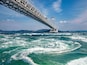 A large suspension bridge over turbulent ocean waters with mountains in the background.