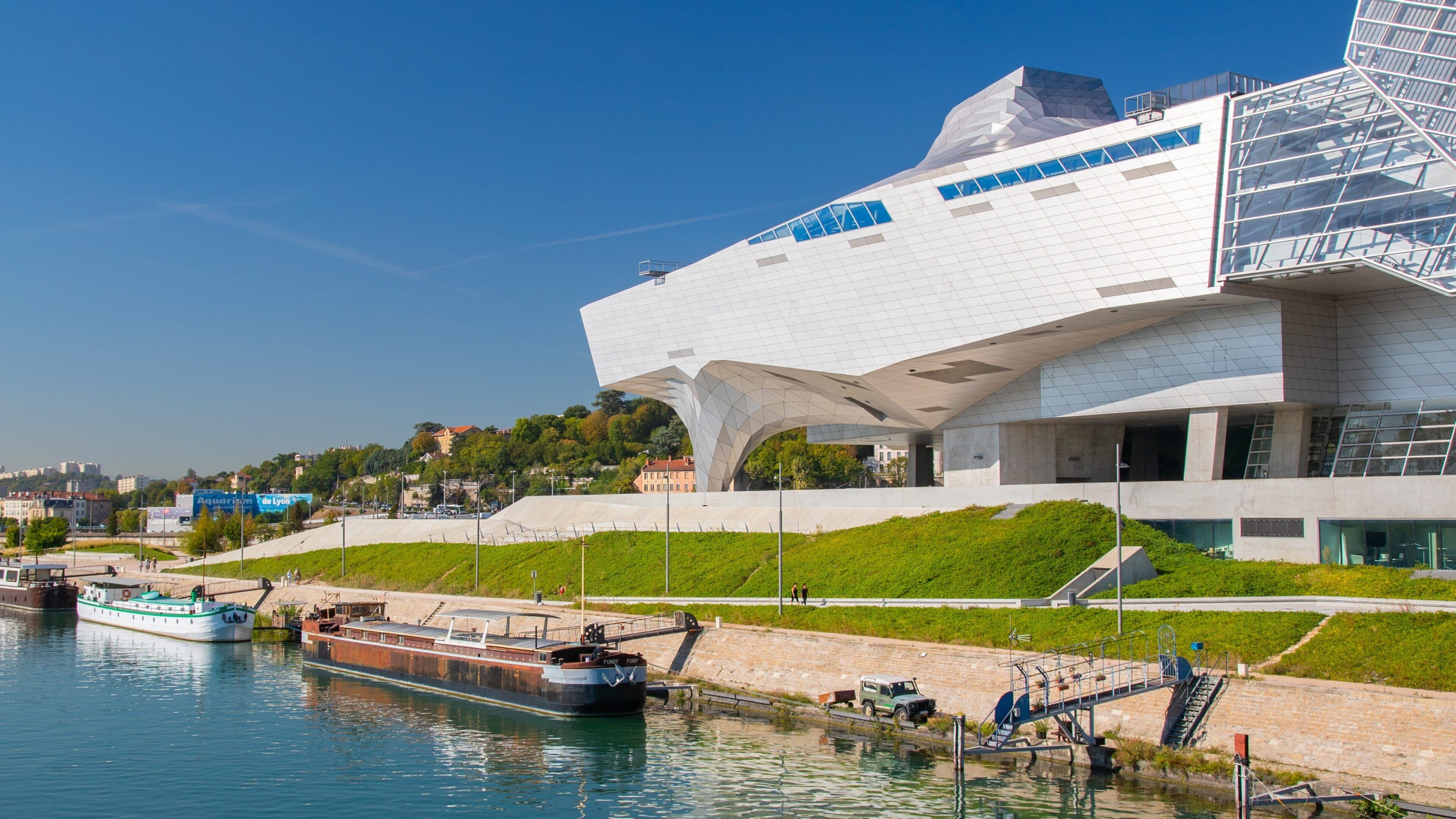 A modern architectural building with a unique design, a river with boats, and a green landscape.