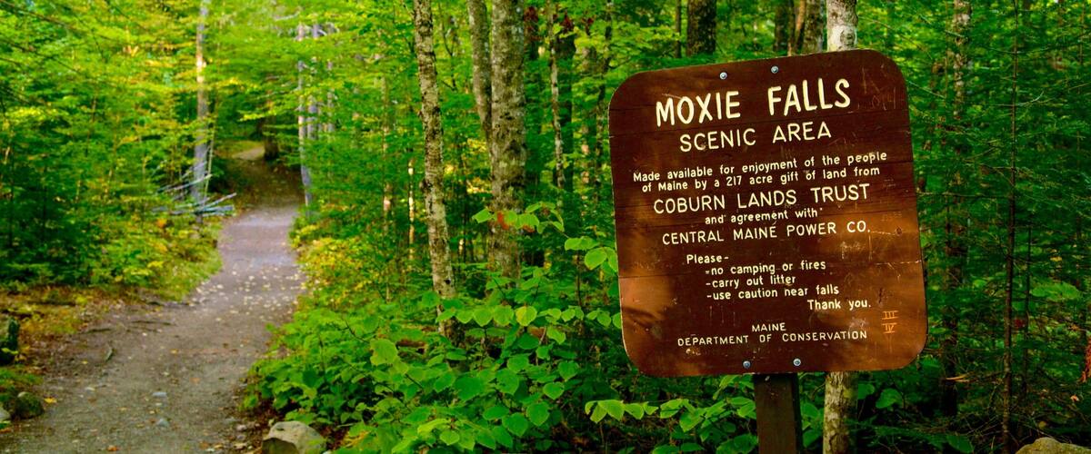 A sign in a forested area labeled 'Moxie Falls Scenic Area' with a path leading through the woods.