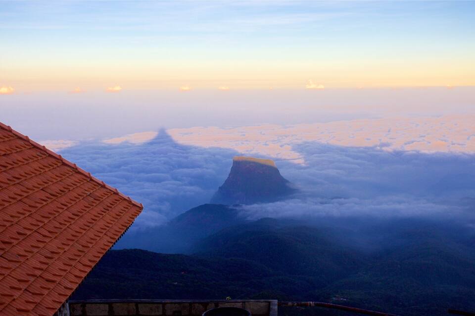 Un pico montañoso sobre las nubes, una edificación de techo rojo y un valle.