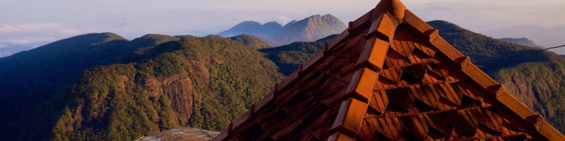 A temple roof with a pointed finial, surrounded by lush green mountains and a clear sky.