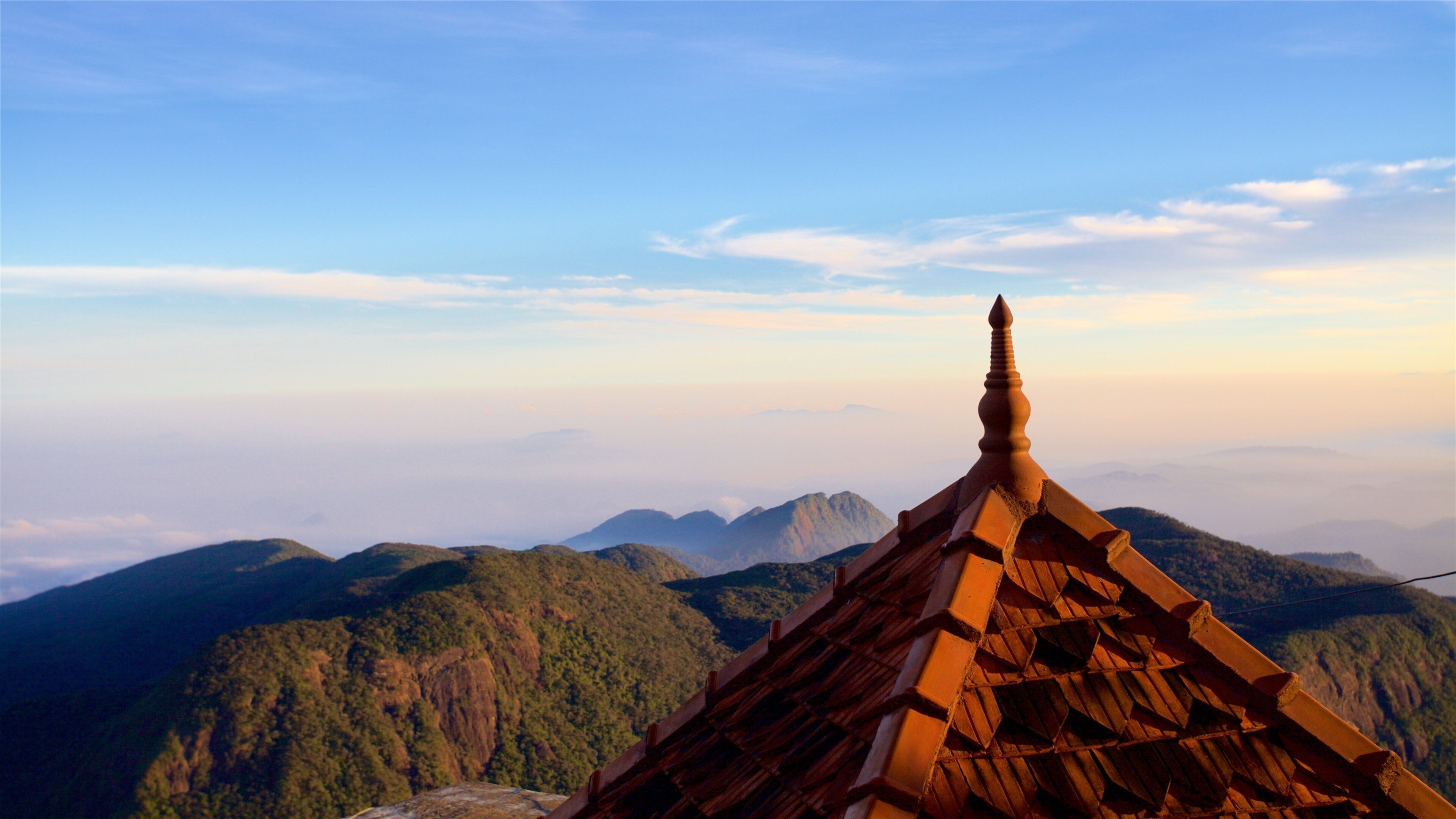 A temple roof with a pointed finial, surrounded by lush green mountains and a clear sky.
