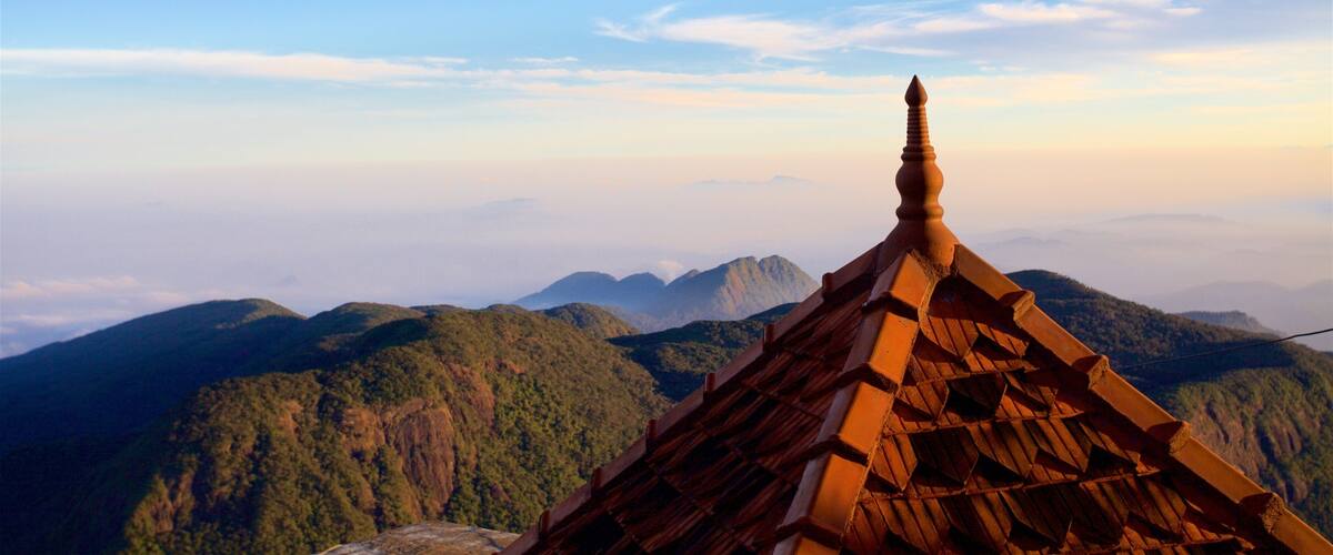 A temple roof with a pointed finial, surrounded by lush green mountains and a clear sky.