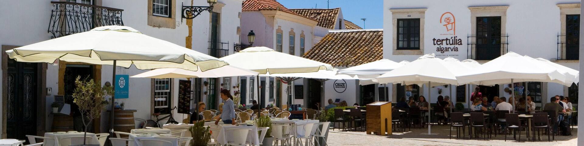 A cobblestone street with outdoor seating and white umbrellas, surrounded by historic buildings with tiled roofs.