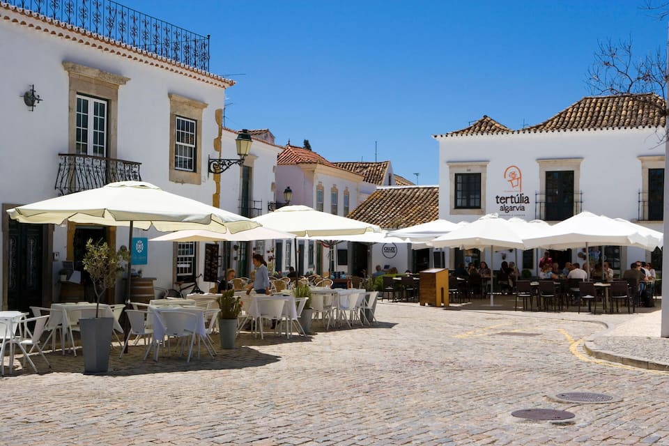 Une rue pavée, bordée de terrasses et ombragée par des parasols blancs, et entourée de bâtiments historiques aux toits de tuiles.
