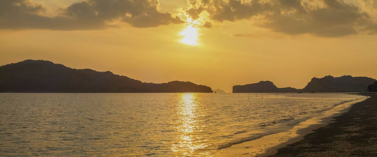 A sunset over a calm sea with a beach and distant mountains.
