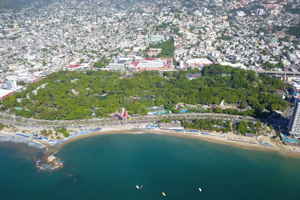 An aerial view of a coastal city with a densely populated urban area, a large park with a flagpole, and a beach with umbrellas.