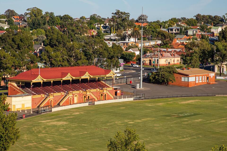 A stadium with a red roof and yellow accents, surrounded by a grassy field and a residential area with houses and trees.