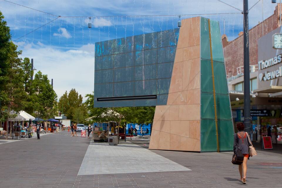 A modern urban square with a large glass building, a Thomas Jewellers store, and a clear blue sky.