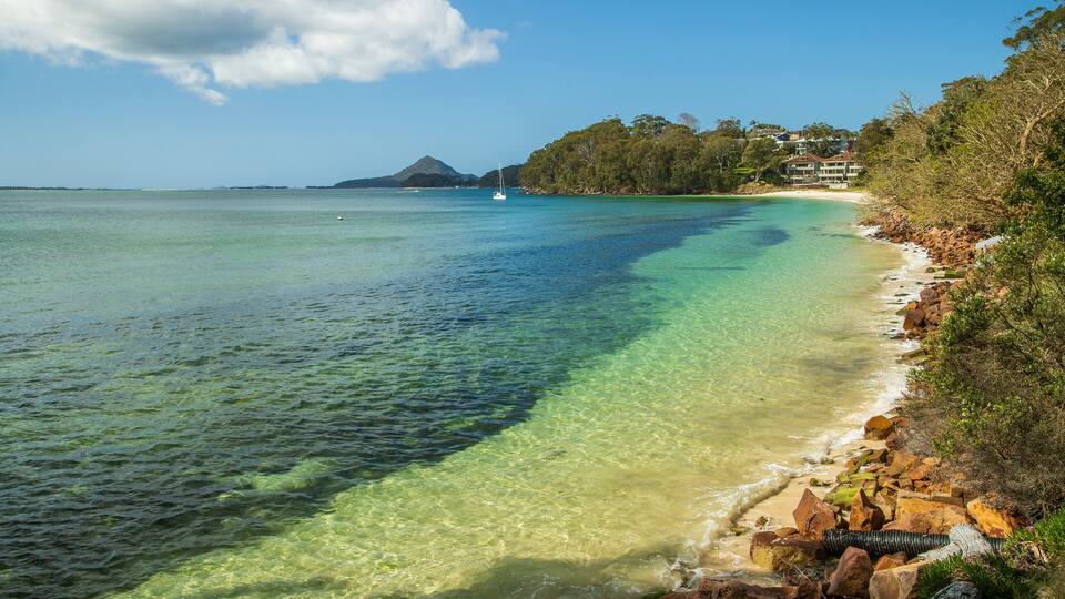 A clear coastal beach with a sailboat, rocky shoreline, and a mountain in the distance.