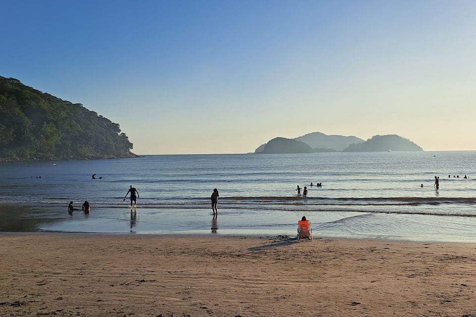 Ein Strand mit badenden und spielenden Menschen, einem Rettungsschwimmerstuhl und einer bewaldeten Klippe im Hintergrund.