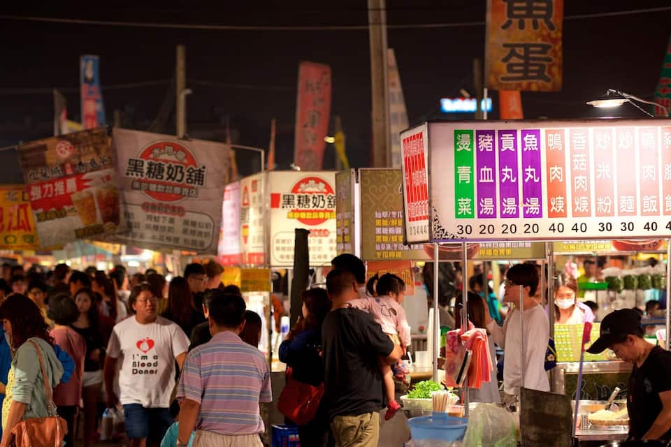 Um mercado de rua movimentado à noite, com diversos quiosques de comida e placas em língua estrangeira.