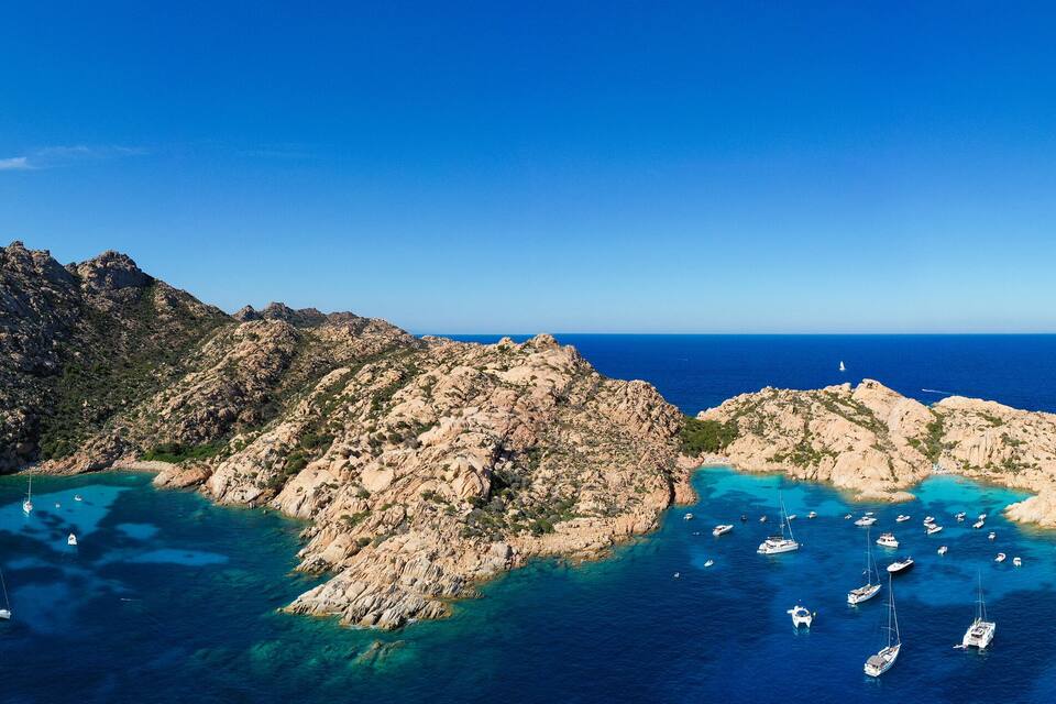 A rocky coastline with clear turquoise waters and numerous sailboats anchored near the shore.