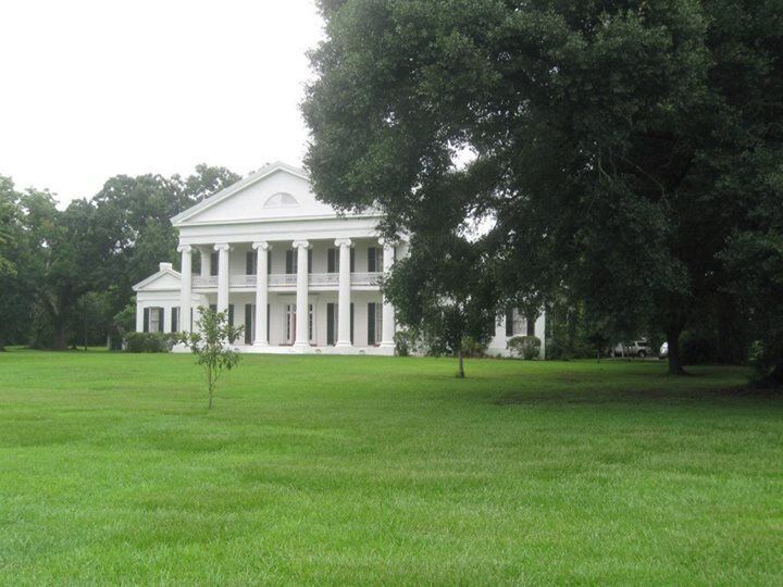 A large white building with columns, surrounded by a grassy field and trees.