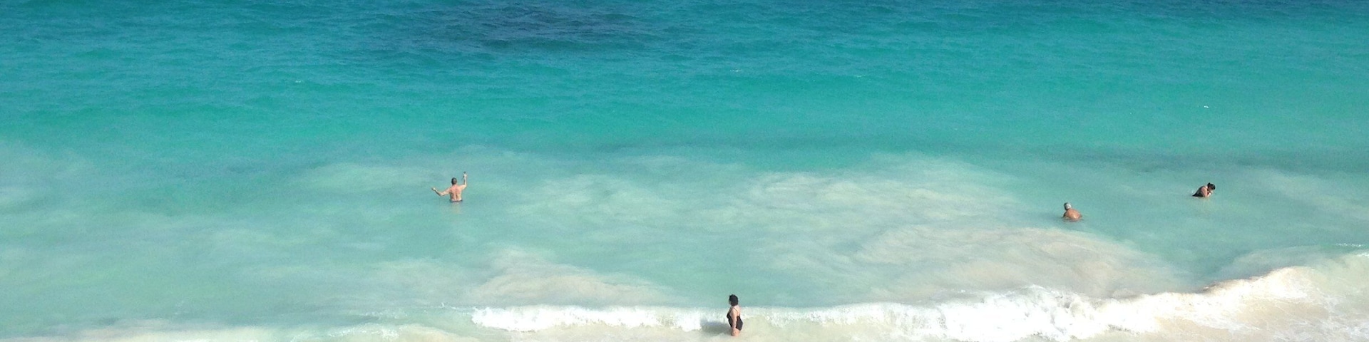 Uma praia com águas turquesa cristalinas, pessoas nadando e caminhando pela orla, e uma cerca de madeira ao primeiro plano.