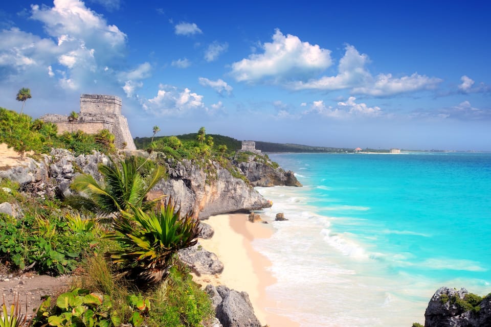 A coastal landscape with a clear blue sky, turquoise waters, and a historic stone structure on a cliff overlooking the beach.