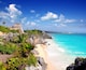 A coastal landscape with a clear blue sky, turquoise waters, and a historic stone structure on a cliff overlooking the beach.