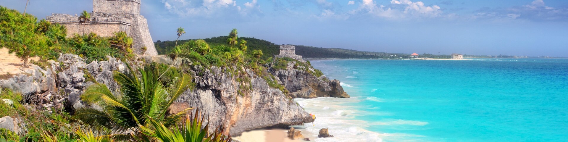 Un paisaje costero con cielo azul despejado, aguas turquesas y una estructura de piedra histórica en un acantilado con vista a la playa.