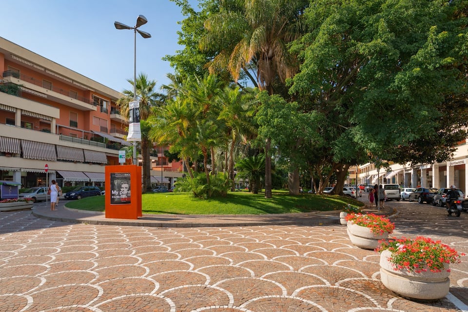 A paved plaza with a patterned design, surrounded by palm trees and buildings.