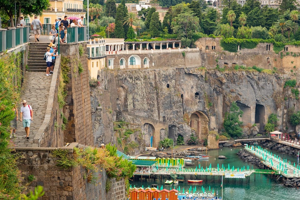 A historic stone fortress with a waterfront promenade, boats, and lush greenery.