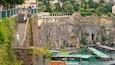 A historic stone fortress with a waterfront promenade, boats, and lush greenery.