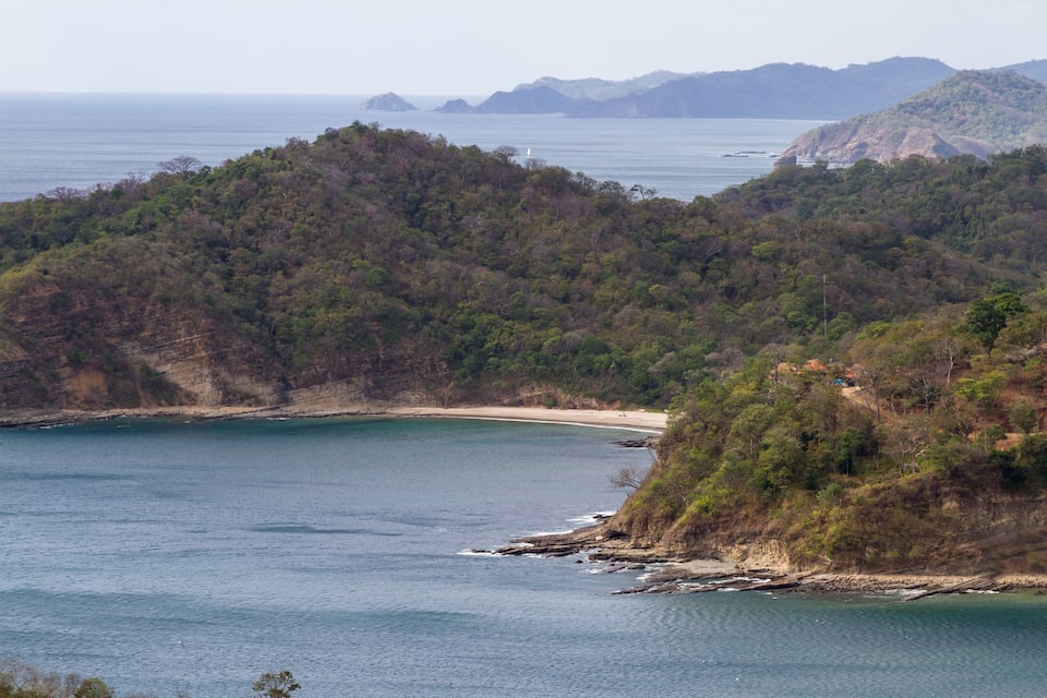 Un paysage côtier avec une plage, une forêt dense et un phare visible au loin.