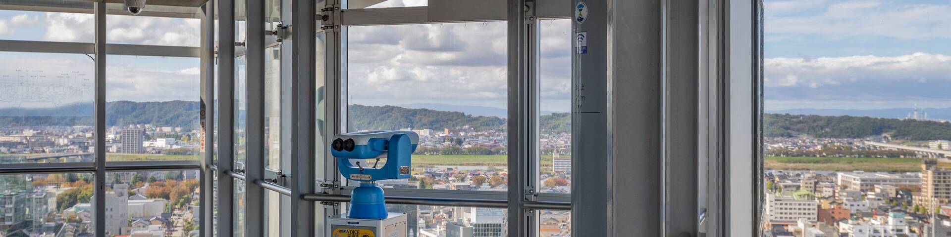 A panoramic view from a high-rise building with large windows, a blue telescope, and a map on the wall.