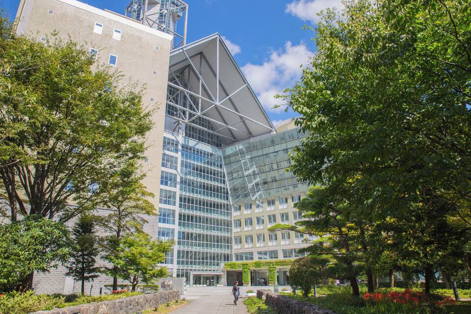 A modern glass building with a unique triangular roof, surrounded by lush greenery and trees.