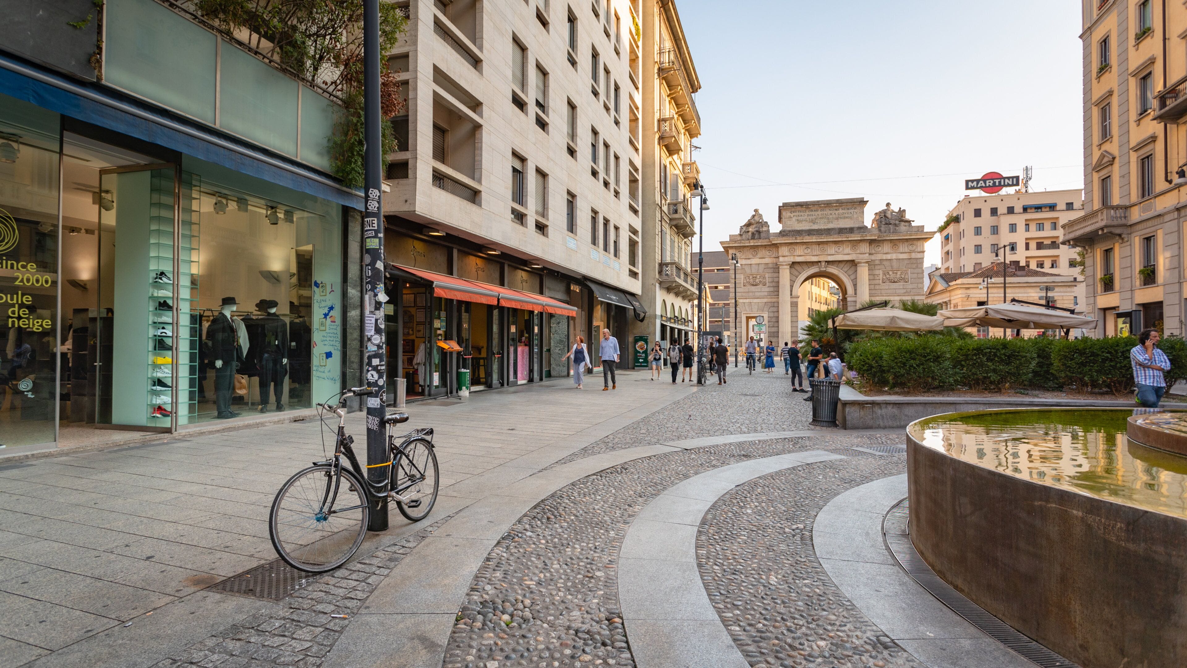 A city square with a fountain, shops, and a historic archway.