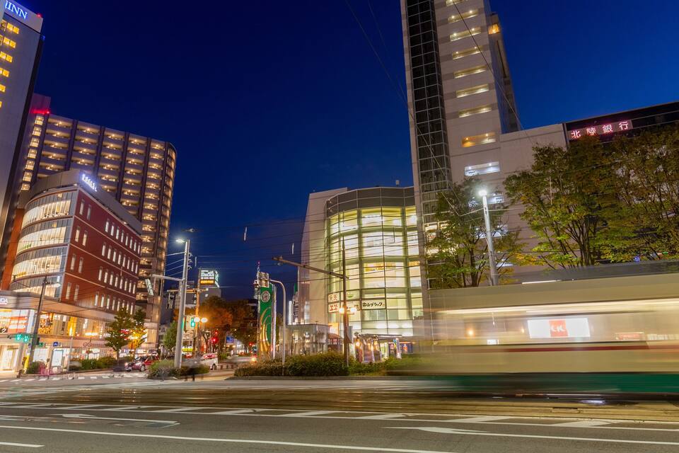 A nighttime cityscape with illuminated buildings, a tram, and a crosswalk.