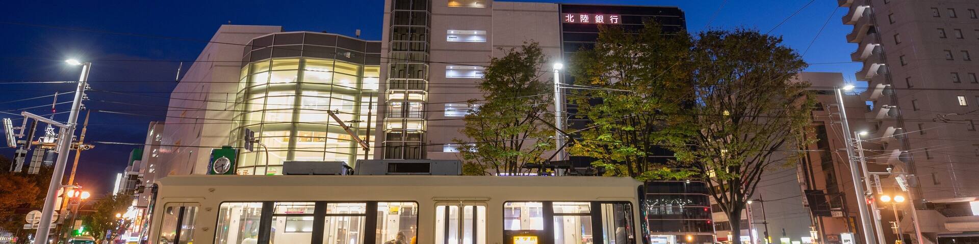 A nighttime cityscape with a tram, a tall building with illuminated windows, and another building with a sign in a foreign language.
