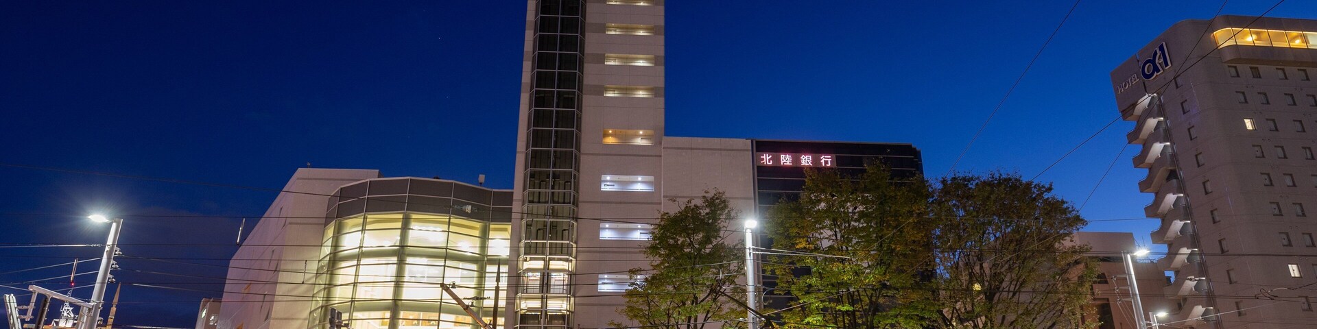 Paisagem urbana noturna com um elétrico, um edifício alto com janelas iluminadas e outro edifício com uma placa em idioma estrangeiro.