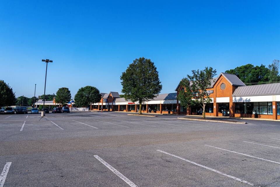 Um parque de estacionamento com vários edifícios, incluindo uma loja com o letreiro 'KOSH', e uma grande árvore ao centro.