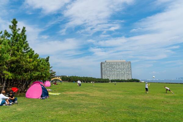 A grassy field with tents, trees, and a large building in the background.