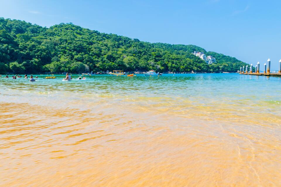 Uma praia com águas cristalinas, pessoas nadando e uma colina arborizada ao fundo.