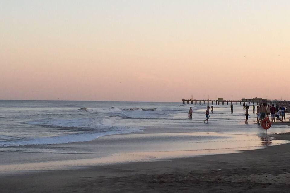 Una platja amb onades al capvespre, un moll al fons i gent passejant pel passeig marítim.
