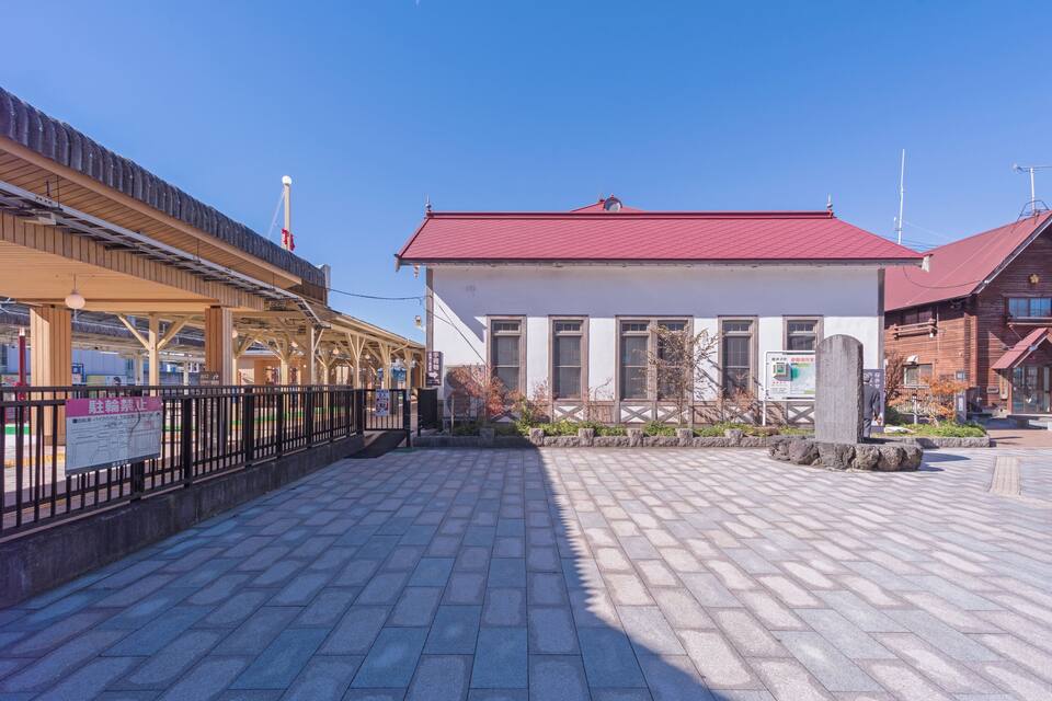 A train station platform with a wooden shelter, a red-roofed building, and stone paving.