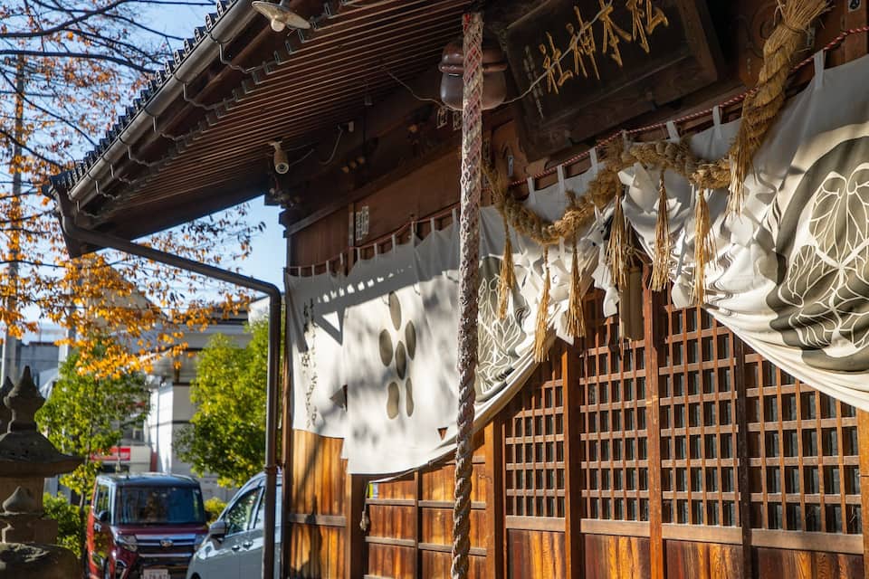 Un edificio tradizionale giapponese con architettura in legno, una corda decorativa pendente e uno striscione bianco con un motivo circolare.