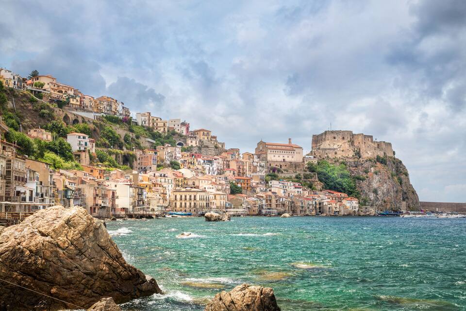 A coastal town with colorful buildings, a rocky shoreline, and a historic fortress on a hill.
