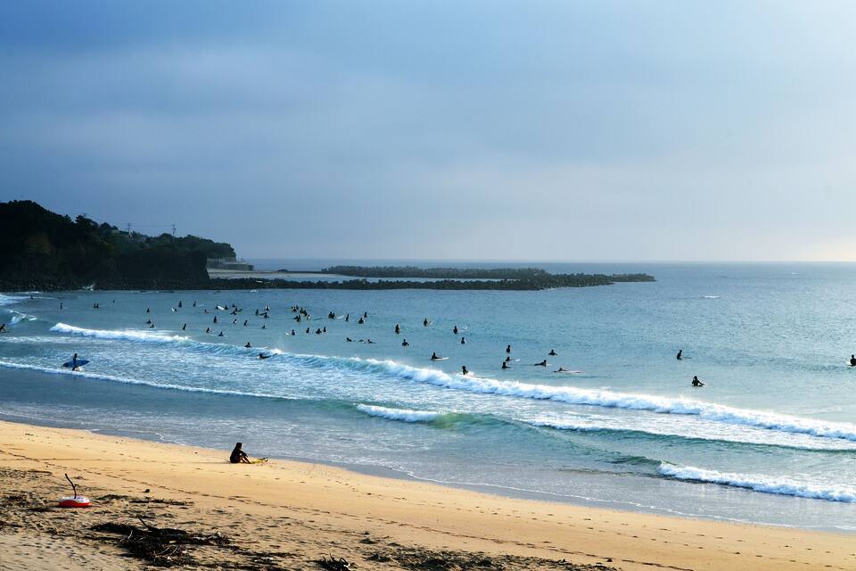 A beach with surfers in the water and a person sitting on the sand.