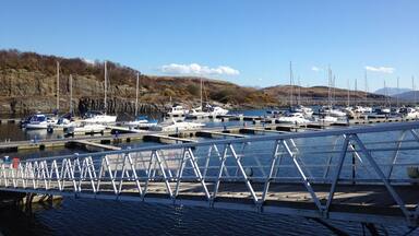 A marina with boats docked, a metal bridge, and hills in the background.