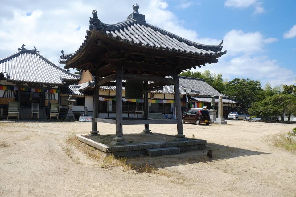 A traditional pavilion with a tiled roof, surrounded by a sandy area with a few parked cars and a statue in the foreground.