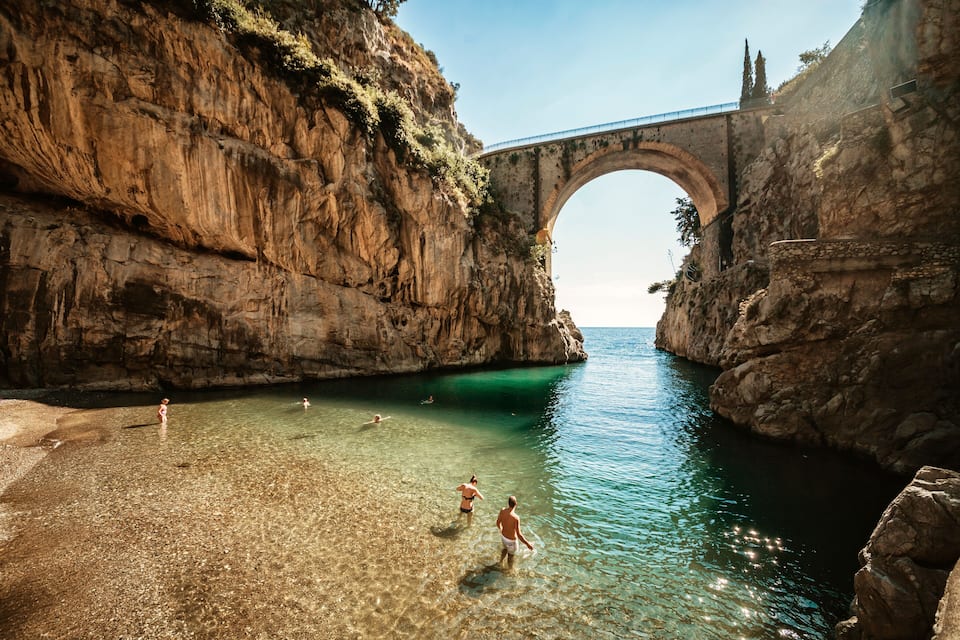 A clear, shallow coastal area with a large rock formation, a bridge, and people swimming.