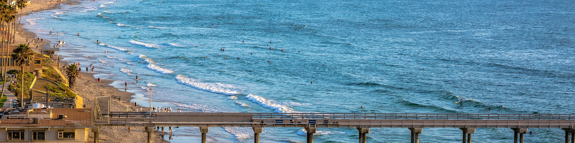 A coastal beach with a pier extending into the ocean, surrounded by residential buildings and palm trees.