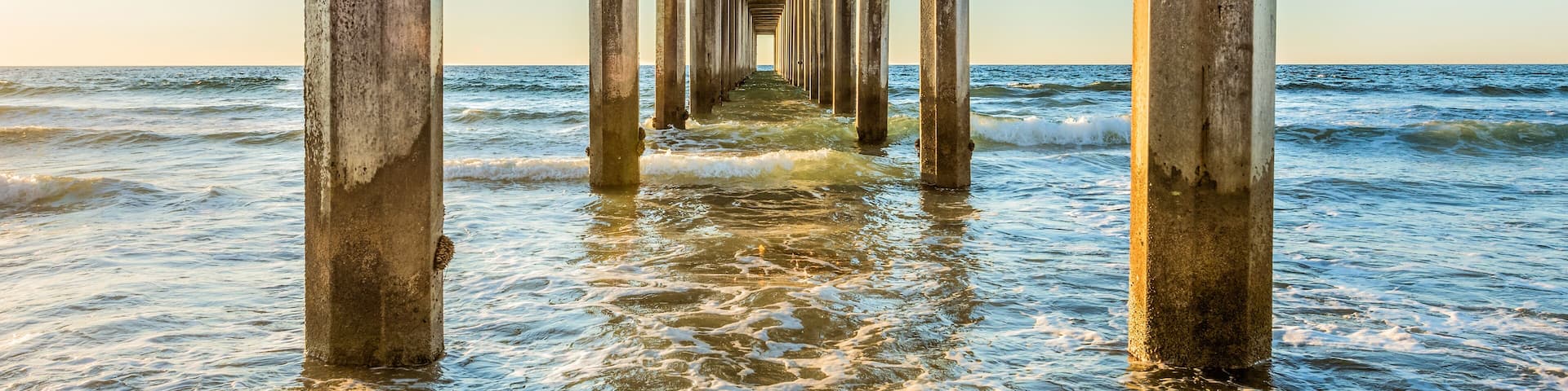 Symmetrical shot under Scripps Pier with poles, pillars, sunny sunlight, sun, blue sky, waves during golden sunset in La Jolla, San Diego, California