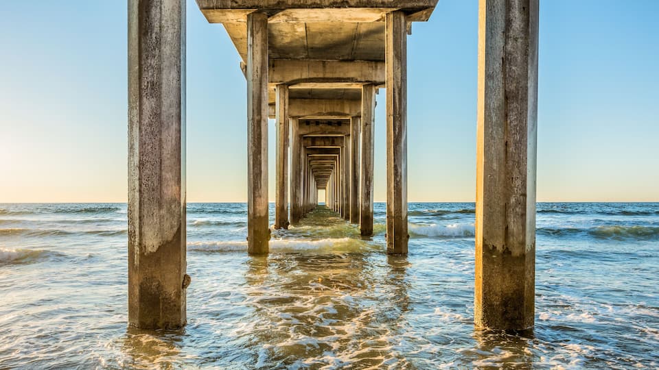 Symmetrical shot under Scripps Pier with poles, pillars, sunny sunlight, sun, blue sky, waves during golden sunset in La Jolla, San Diego, California